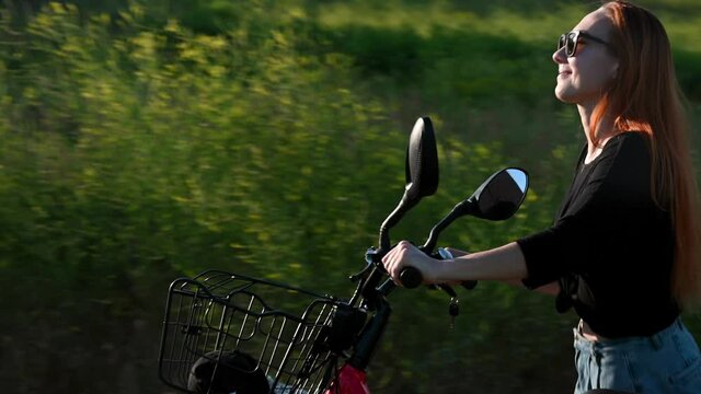 Beautiful caucasian girl at background of green grass pushing uphill red electro bicycle at sunny summer day. lighting effects, sun glare. fitness, people, healthy lifestyle concept