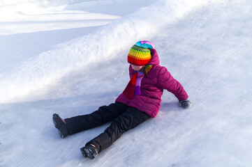 Fototapeta na wymiar Child rolls down an ice slide. Bright clothes rainbow hat.