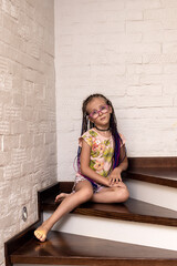 a charming European girl of six with African colored pigtails and glasses is sitting on the steps of her house