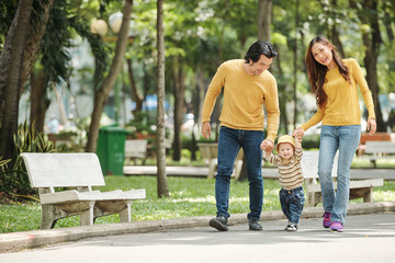 Joyful Vietnamese husband, wife and baby boy in jeans and sweaters walking in local park on sunny day