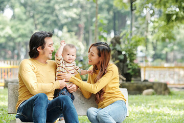 Happy young Vietnamese man and woman sitting on bench in park and adjusting sweater of thier little...