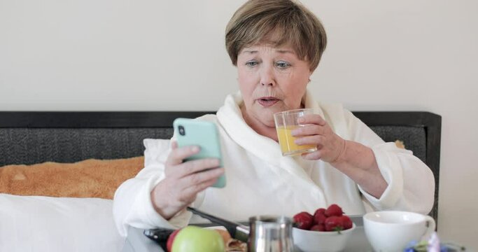Elderly beautiful lady making shocked facial expession while using her smartphone. Mature woman in home rob having breakfast and drinking juice while getting bed news on her phone.
