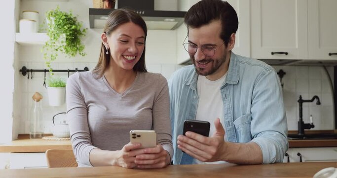 Cheerful couple sit at dining table in kitchen hold smart phones having fun use on-line services, show photos, share new cool app, laughing enjoy modern tech usage, virtual real communication concept