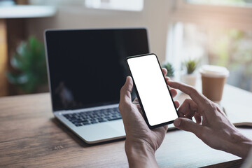 business man holding mockup smartphone with blank white screen and computer laptop on wooden table in cafe shop