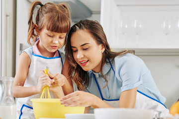 Positive young woman teaching daughter how to whisk eggs with sugar when making cake frosting