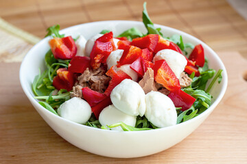 Fresh summer vegetables and green herbs, red pepper, arugula, canned tuna and mozzarella as a salad in the white bowl on wooden background.