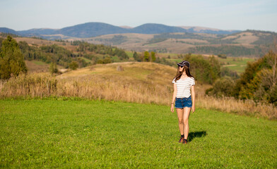 Naklejka na ściany i meble Carefree happy woman in sunglasses, cap and jeans shorts walk on green grass meadow on top of mountain enjoying nature. Freedom