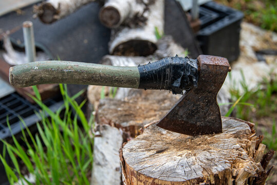 An Old Rusty Ax With A Wooden Handle Is Rewound With A Black Ribbon, Stuck In A Birch Stump Against A Background Of Green Grass And Birch Firewood. Chopping.