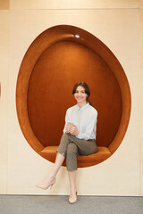 Full length portrait of smiling young businesswoman looking at camera while sitting in egg-shaped...