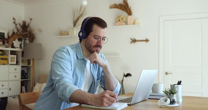 Millennial man sit at desk in living room looks at laptop screen listen audio through headphones use exercise book makes notes learn language develop improve gain new knowledge. Self-education concept