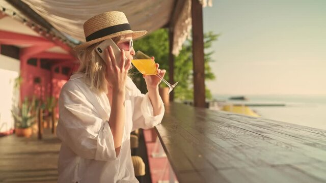 An attractive young blonde woman wearing sunglasses and hat is talking on the smartphone while sitting in the cafe on the beach in the morning