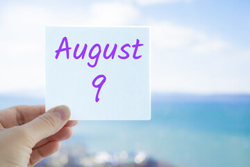 August 9th. Hand holding sticker with text August 9 on the blurred background of the sea and sky. Copy space for text. Month in calendar concept