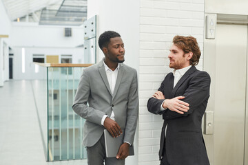 Waist up portrait of two successful businessmen wearing suits chatting while standing in while office building interior, copy space