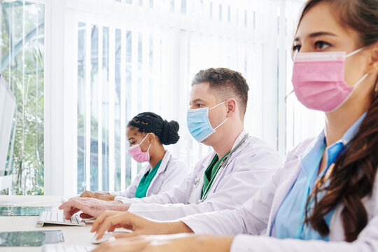 Young doctors in medical masks working on computer in laboratory and entering patients data and results of massive research of new vaccine