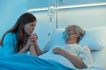 Worried daughter or granddaughter holding the hand of her mother in hospital as the elderly woman lies asleep or in a coma on a bed wearing a positive oxygen mask to assist breathing - 366060874