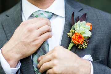 orange flower and green succulent boutonniere on groom suite while he is adjusting his tie for...