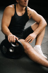 Fototapeta na wymiar Close-up of Muscular athletic man n with perfect beautiful body wearing sportswear sitting on the gym floor with weight from the barbell after heavy workout training in dark modern gym.