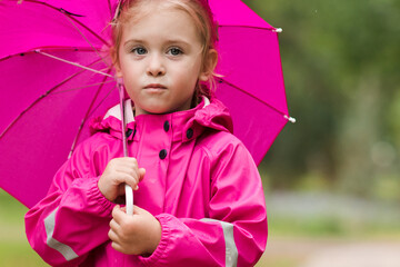 Cute child girl 2-3 year old wearing pink stylish raincoat holding umbrella standing in park over green nature background. Looking at camera. Childhood. Autumn season.