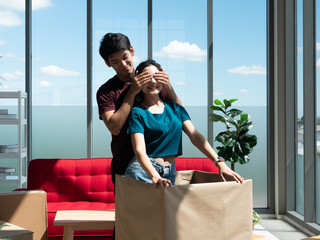 Asian young boy surprise pretty girl who standing inside big cardboard by close her eyes from relocation and moving to new home while smiling and enjoying open stuff box in living room romantic scene