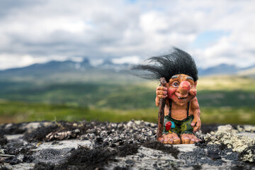 Funny norwegian troll figure with big nose and walking stick outdoors in the mountains. Hair standing up because of windy conditions. - obrazy, fototapety, plakaty