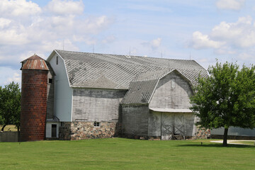 a large old stone foundation white barn and stone foundation and brick silo with a bright green grass lawn and shade trees
