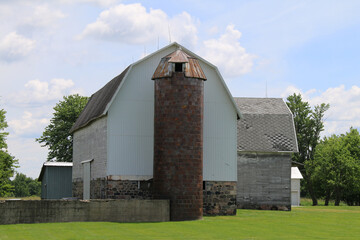 a large white farm barn and stone foundation and silo with green grass lawn and trees