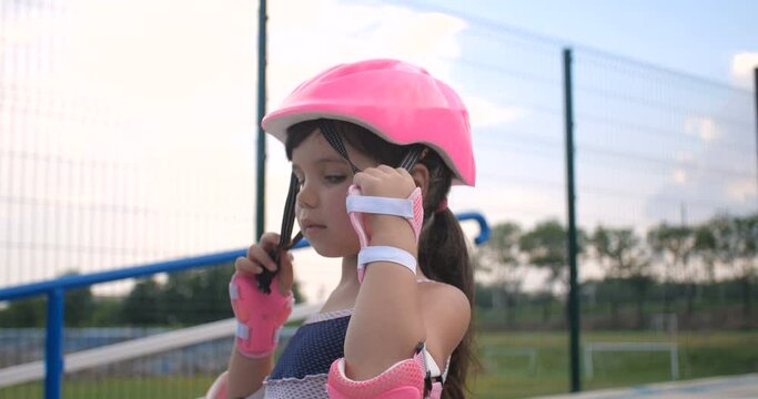 A little girl first time came to the roller park to learn roller skating.  Close up portrait of a pretty child of preschool age putting on a pink safety helmet, 4K slow motion 50 FPS 