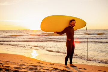 Fototapeta na wymiar Young bearded surfer in wetsuit with yellow surfing longboard on a ocean coast at sunset. Water sport adventure camp and extreme swim on summer vacation.