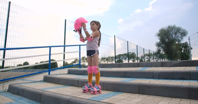 A little girl first time came to the roller park to learn roller skating. Pretty child of preschool age putting on a bright pink safety helmet, 4K slow motion 50 FPS 