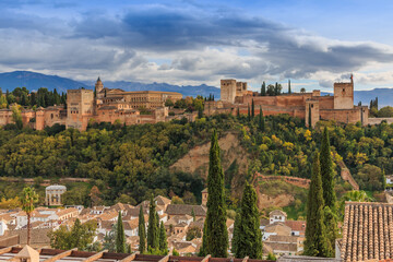 Fototapeta na wymiar Spanish city of Granada in Andalusia with clouds and mountains in the background. Historic Alhambra fortress with trees and buildings on a hill. Red tile roofs of the buildings to the autumn mood
