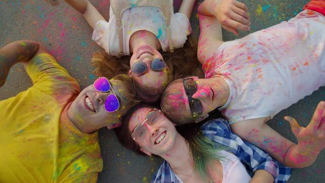 A multi-ethnic group of friends, smeared with Holi powder, lies on the pavement, laughing, looking up and gesturing at the camera