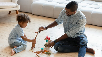 Loving african American young father sit on warm wooden floor play toys bricks cubes with small...