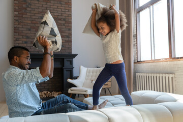 Overjoyed young african American dad and little daughter engaged in pillow fight in living room,...
