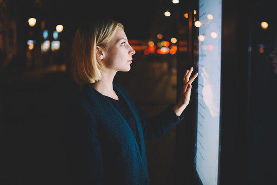 Attractive young woman consults on modern big timetable outdoors, female caucasian tourist touching information kiosk digital screen while standing at night city with beautiful lights on background