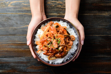 Stir fried kimchi with pork on cooked rice in a bowl holding by hand on wooden background, Korean...
