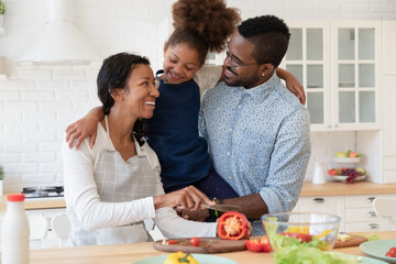 Overjoyed young african American couple parents have fun cook healthy breakfast or salad with smiling little ethnic daughter, happy biracial family mom and dad enjoy preparing food with girl child