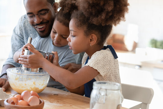 Loving african american father make dough making tasty sweet breakfast pastry with excited little children, happy biracial dad and small kids cook prepare pancakes cookies pie in kitchen together
