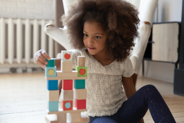 Playful little african American girl sit on warm floor at home play construct tower with wooden...