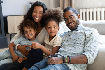Portrait of smiling young african American family with two small kids sit relax on couch in living room together, happy biracial mom and dad hug cuddle with little children at home, show love care - Powered by Adobe