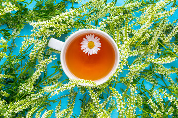 Obraz na płótnie Canvas A cup of tea with chamomile on a blue background decorated with green wildflowers. Copy space. Summer composition.