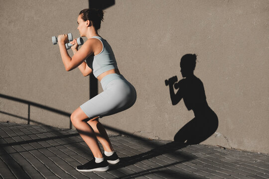 Athletic girl crouches with dumbbells. Sporty fitness woman in leggings and top outdoors doing leg workout