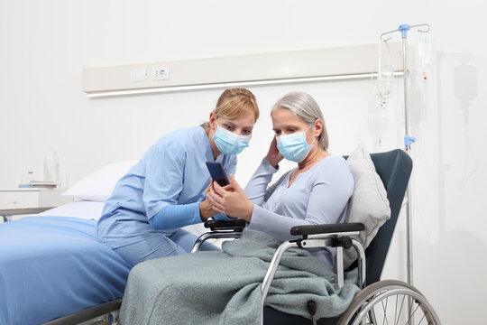 Nurse Helps With Cell Phone To Contact The Elderly Lady's Family In The Wheelchair, Wearing Surgical Protective Medical Masks In Hospital Room, Concept Of Isolation From Corona Virus Covid 19