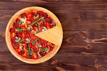 Pizza hot with spicy salami, arugula, cherry tomatoes, mushrooms and texas spice mix, without one slice on a wood platter which is on wooden rustic table, top view