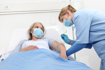 nurse with the syringe injects the vaccine to the elderly woman patient lying in the hospital room bed, wearing protective gloves and medical surgical mask, coronavirus covid 19 protection concept