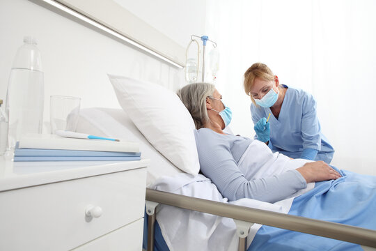 Nurse With The Syringe Injects The Vaccine To The Elderly Woman Patient Lying In The Hospital Room Bed, Wearing Protective Gloves And Medical Surgical Mask, Coronavirus Covid 19 Protection Concept