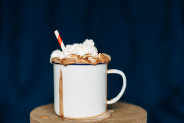 Enamel mug with chocolate milk, whipped cream and a straw in front of a dark blue background