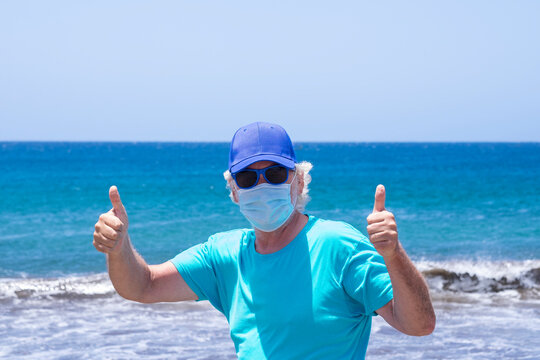 Front portrait of a senior bearded man with white hair at the beach gesturing ok with fingers, wearing a mask because of the coronavirus, horizon over water - concept of active seniors on vacation