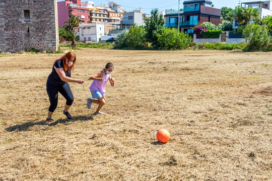 Mother and daughter playing soccer with an orange ball in a field with grass wearing masks to prevent coronavirus