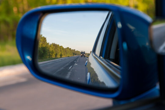 Rear view in the side mirror of a blue sedan with the reflection of a gray truck on an asphalt road on a summer day with green trees on the sides of the highway.