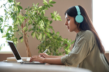 Indian teen girl school college student wear headphones learning watching online webinar class looking at laptop computer elearning lesson making notes or video calling virtual meeting remote teacher.